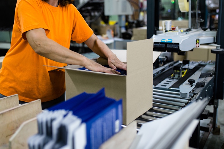 A close-up of a worker in an orange shirt placing things in a box on a conveyor belt line next to machinery.