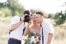 A wedding photographer showing a camera preview to a smiling bride and groom standing closely outdoors in soft light.