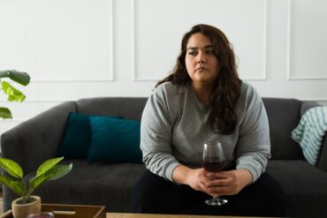 A woman with a sad look on her face sits on a dark-gray couch holding a wine glass with red wine in it.