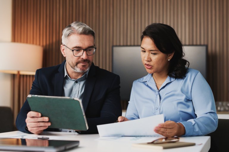 A man and a woman sit at a table looking at a piece of paper the woman is holding. The man is holding a tablet.