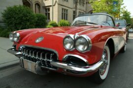Red vintage convertible car with chrome details parked on street, buildings and greenery reflected on glossy surface.