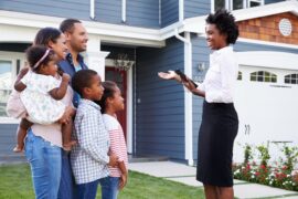 A realtor talking with a family of five on the lawn in front of a blue two-story home. Everyone is smiling.