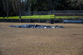 A large, round black tarp covers the pitcher's mound in the middle of an empty baseball field. Heavy objects sit on the tarp.