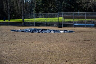 A large, round black tarp covers the pitcher's mound in the middle of an empty baseball field. Heavy objects sit on the tarp.