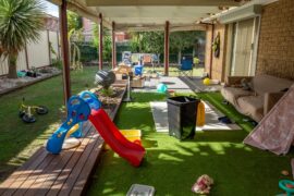 A messy backyard play area with artificial grass, a toddler slide, and scattered toys on a covered patio.