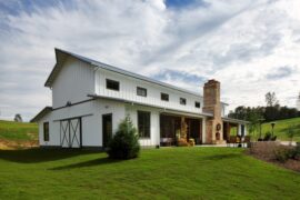 A white ranch farmhouse with a front lawn and intricate landscaping under a cloudy sky in a rural setting.
