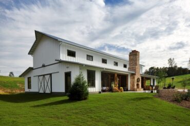 A white ranch farmhouse with a front lawn and intricate landscaping under a cloudy sky in a rural setting.