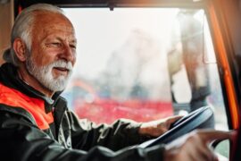 A senior man sitting in the driver's seat of a truck cab, one hand on the wheel, the other tapping on a GPS.