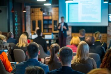 A lecturer speaking to an audience at a professional workshop with attendees sitting and listening indoors.