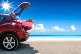 a red-colored van parked on a sandy beach near blue water while reflecting off sunlight under a clear, blue sky.