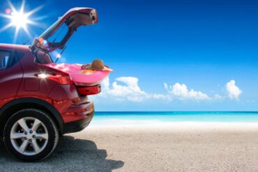 a red-colored van parked on a sandy beach near blue water while reflecting off sunlight under a clear, blue sky.