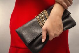 A woman wearing a red dress holds a black leather clutch bag with gold chains wrapped around it.