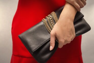 A woman wearing a red dress holds a black leather clutch bag with gold chains wrapped around it.