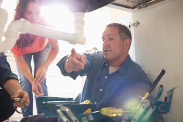 A man points to the plumbing beneath a sink while a woman leans in for a closer look. A light shines in the background.