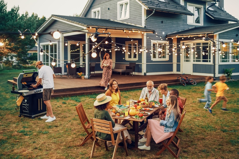 A large family with grandparents, parents, and children spending time in the home backyard around a table.