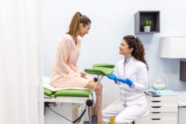 A female patient sits on a green exam table talking to a female healthcare professional who sits on a stool.