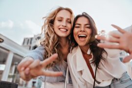 Two smiling women lean toward the camera outdoors, one flashing a peace sign and the other reaching forward.