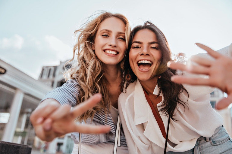 Two smiling women lean toward the camera outdoors, one flashing a peace sign and the other reaching forward.