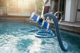 A man cleaning a home swimming pool with a vacuum that is attached to a long blue tube. The vacuum is out of the water.