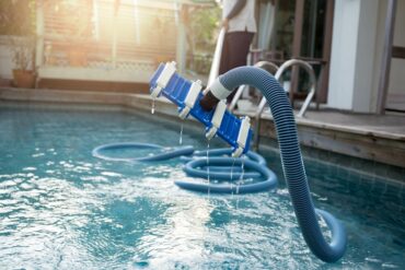 A man cleaning a home swimming pool with a vacuum that is attached to a long blue tube. The vacuum is out of the water.