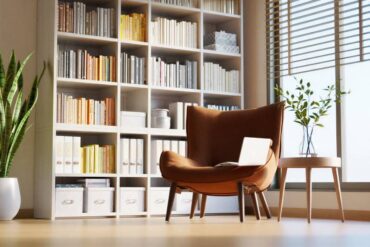 Cozy home library with white bookshelves, a brown lounge chair, laptop, potted plants, and bright window light.