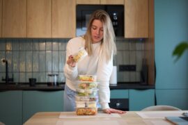 Woman opens a food container while arranging stacked prepared meals on a table in a bright modern kitchen.