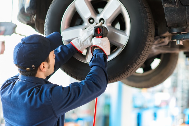 A mechanic wearing white gloves using a small drill to remove a tire from a car in an auto repair shop.