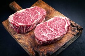 Two raw Wagyu ribeye steaks display rich marbling on a rustic wooden cutting board against a dark background.
