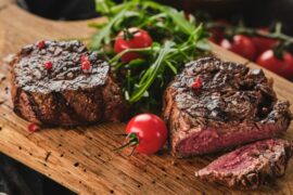 A wooden board holding grilled steak pieces alongside cherry tomatoes, some on the vine and some off, plus leafy greens..