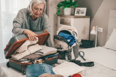 An older woman closes a suitcase filled with clothing that lies on her bed. A backup with more items rests next to it.