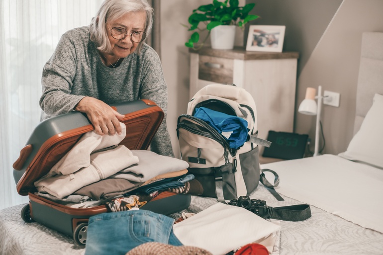 An older woman closes a suitcase filled with clothing that lies on her bed. A backup with more items rests next to it.