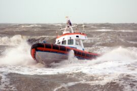 A white and orange rescue boat is being tossed around by high waves on a cloudy day. The waves are splashing against the boat.
