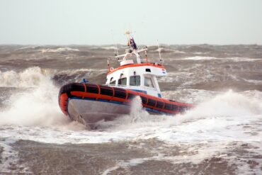 A white and orange rescue boat is being tossed around by high waves on a cloudy day. The waves are splashing against the boat.