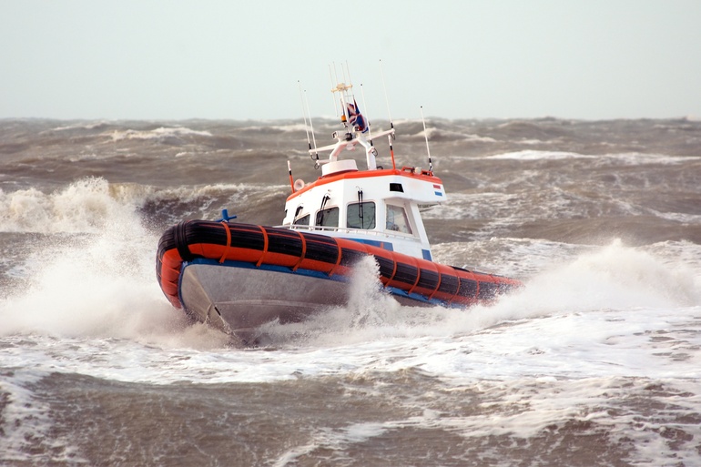 A white and orange rescue boat is being tossed around by high waves on a cloudy day. The waves are splashing against the boat.