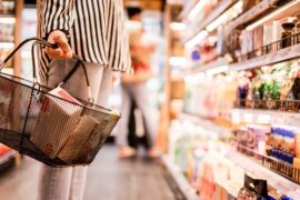 A woman is standing next to a row of merchandise. She’s holding a basket in her hand that is filled with items.