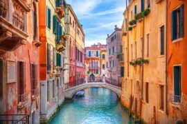 A colorful Venetian canal lined with historic buildings and a stone bridge spanning turquoise water under a bright sky.