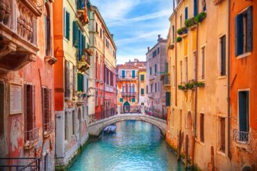 A colorful Venetian canal lined with historic buildings and a stone bridge spanning turquoise water under a bright sky.
