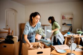 Two women are in a room. The one in the foreground is packing stuff into a cardboard box.