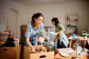 Two women are in a room. The one in the foreground is packing stuff into a cardboard box.