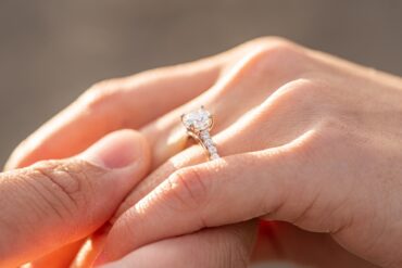 A close-up of a diamond engagement ring on a person's finger as hands gently touch, illuminated by warm sunlight.