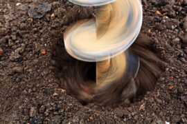 A close-up view of a spinning drill bit boring into dark soil, with loose dirt moving around the hole.