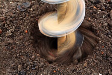 A close-up view of a spinning drill bit boring into dark soil, with loose dirt moving around the hole.
