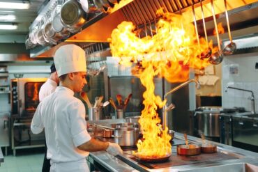 Chef cooking on a stovetop in a brightly lit commercial kitchen. A large orange flame rises from the pan.