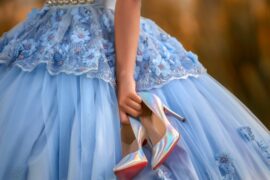 Person in a light blue floral quinceañera gown holds shimmering iridescent high heels beside the full tulle skirt.