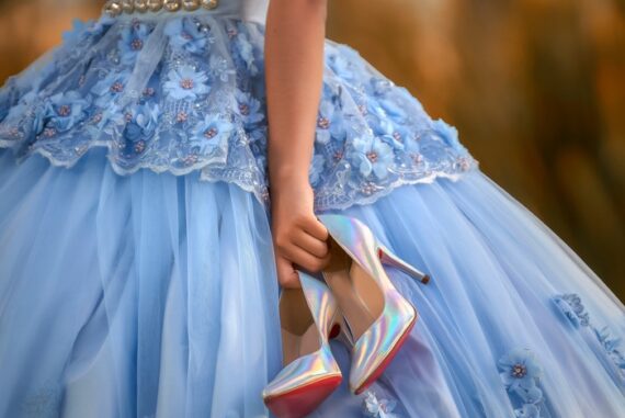 Person in a light blue floral quinceañera gown holds shimmering iridescent high heels beside the full tulle skirt.