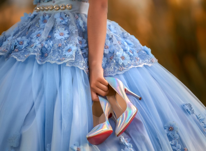 Person in a light blue floral quinceañera gown holds shimmering iridescent high heels beside the full tulle skirt.