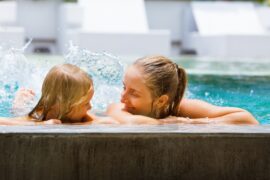 A close-up of a mother and her young daughter holding onto the edge of a pool, splashing while smiling at each other.
