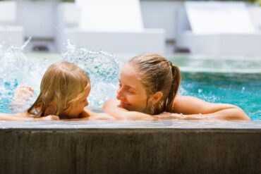 A close-up of a mother and her young daughter holding onto the edge of a pool, splashing while smiling at each other.