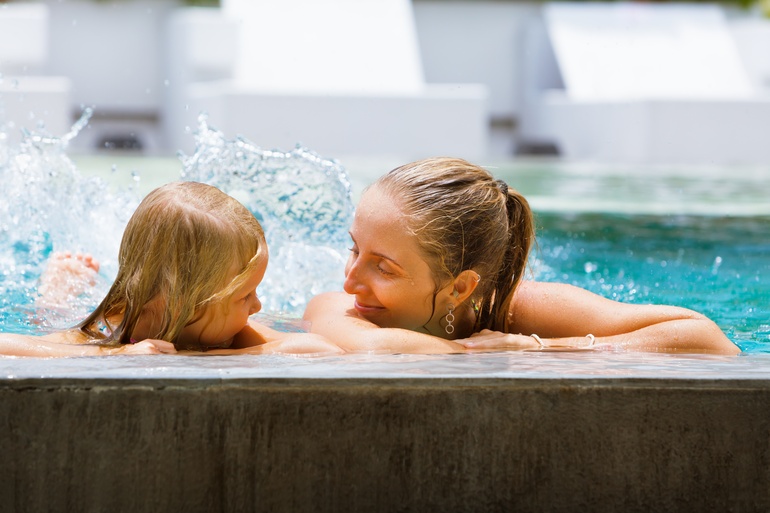 A close-up of a mother and her young daughter holding onto the edge of a pool, splashing while smiling at each other.