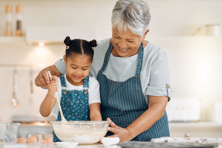 Grandmother smiles while helping her young granddaughter stir batter in a mixing bowl together in a bright home kitchen.